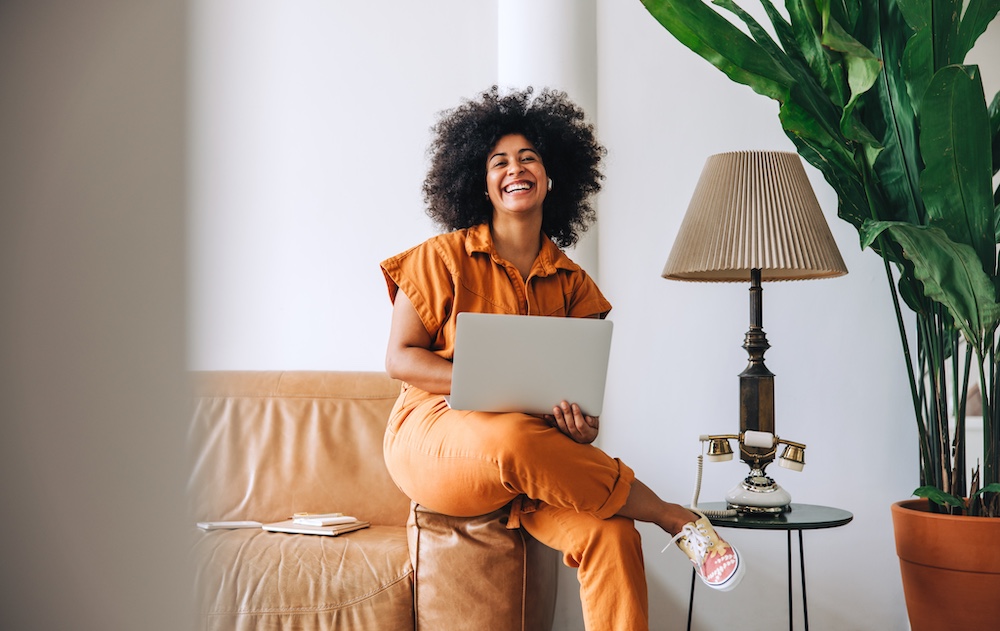 Businesswoman smiling cheerfully in an office lobby Businesswoman with Afro hair smiling cheerfully in an office lobby. Happy young businesswoman holding a laptop while sitting on a couch in a modern workplace.